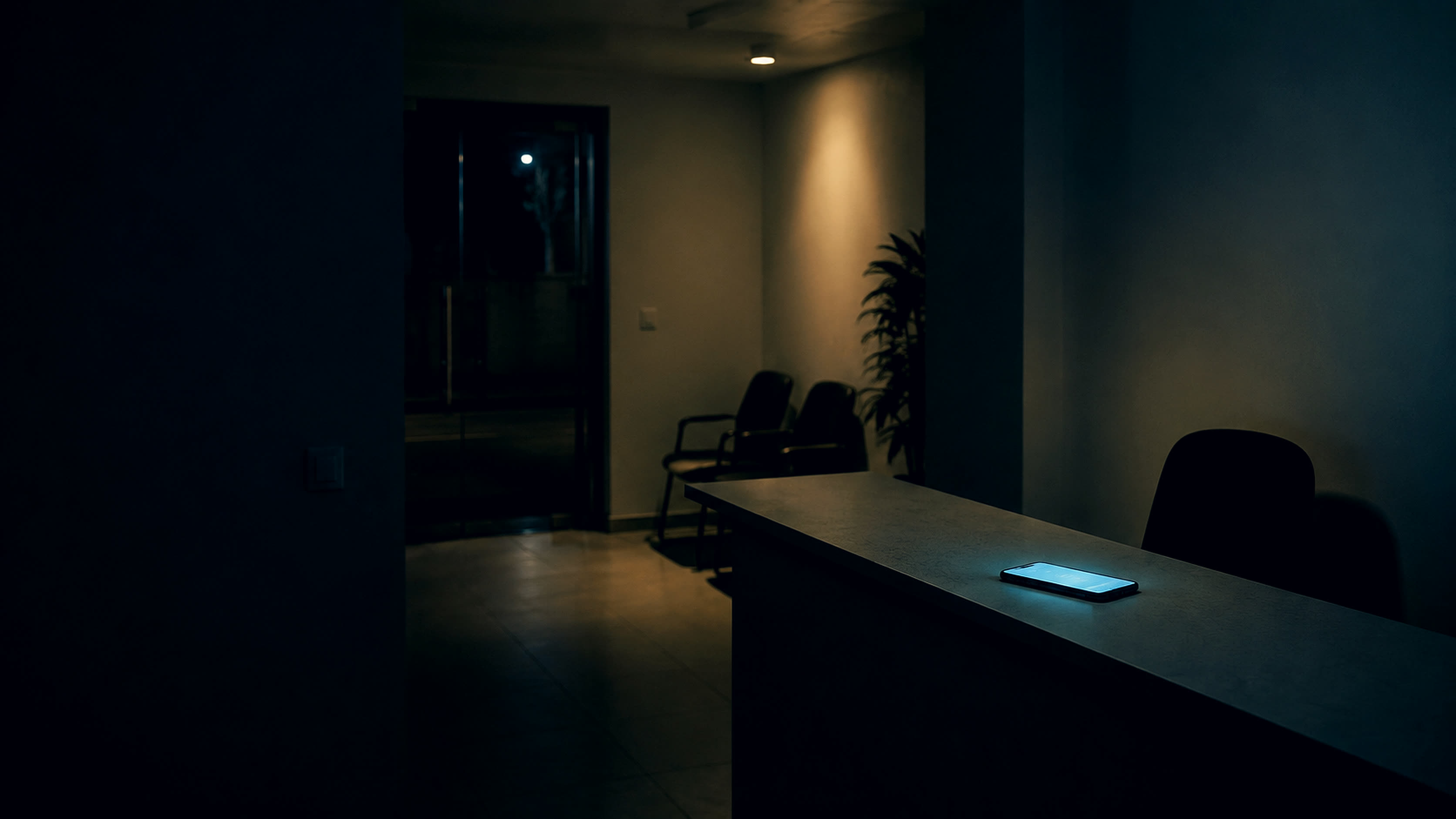 Recepção vazia de uma empresa pequena às sete da noite: balcão sem ninguém, cadeiras de espera vazias e um celular aceso sozinho sobre a bancada.
